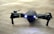 A sleek black drone with extended propeller arms and illuminated front lights is positioned on a soft beige surface. The drone has markings in yellow and is detailed with inscriptions on one arm.
