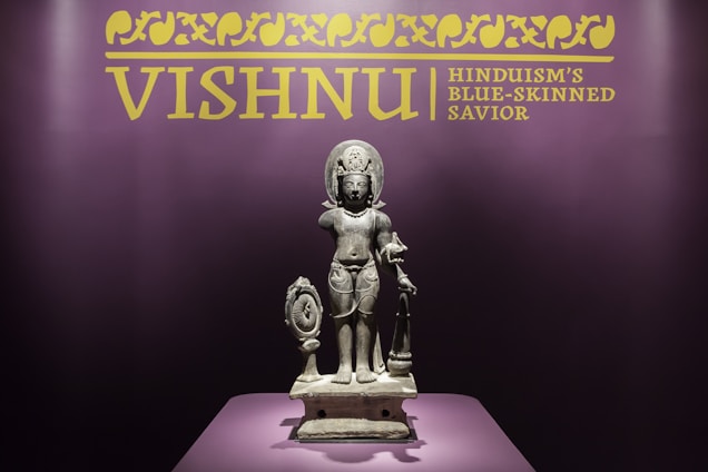 A stone statue of a deity stands centrally on a pedestal, surrounded by a dark purple backdrop. Above the statue, there is a yellow sign with ornate patterns and text that reads 'VISHNU Hinduism's Blue-Skinned Savior'. The statue depicts a finely detailed figure with traditional adornments and holds various items in its hands.