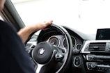 A person is seated inside a BMW car, grasping the steering wheel with their left hand. The dashboard is prominently visible with various controls, including an air conditioning panel, start/stop button, and a digital display. The steering wheel features the BMW logo in the center.