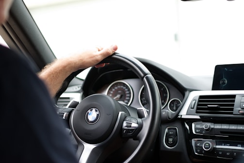 A person is seated inside a BMW car, grasping the steering wheel with their left hand. The dashboard is prominently visible with various controls, including an air conditioning panel, start/stop button, and a digital display. The steering wheel features the BMW logo in the center.