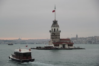 A historic tower stands on a small island in the water, with a Turkish flag flying at the top. Boats navigate the water surrounding the tower. In the background, the skyline of a city is visible under an overcast sky.