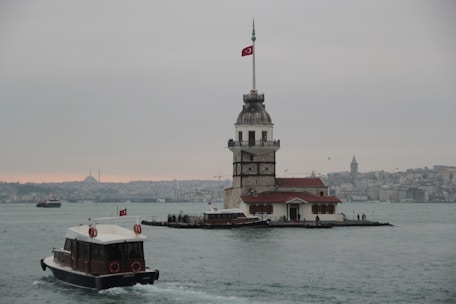 A historic tower stands on a small island in the water, with a Turkish flag flying at the top. Boats navigate the water surrounding the tower. In the background, the skyline of a city is visible under an overcast sky.