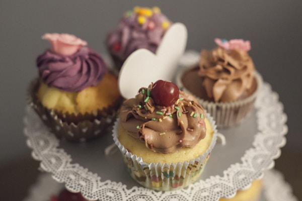 Four decorated cupcakes are arranged on a lace doily-covered cake stand. The cupcakes are topped with various frosting styles and decorative elements, including a pink flower, colorful sprinkles, and a cherry. The background is softly blurred, keeping focus on the cupcakes.