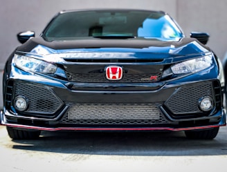A sleek black car with a prominent front grille and distinctive red Honda emblem. The headlights are sharp and modern, with a sporty bumper design featuring mesh patterns and fog lights. The gloss of the black paint reflects light, emphasizing the car's polished finish.