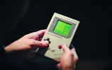 A person is holding a classic handheld gaming device with a green screen displaying a video game. The device appears to be a Game Boy with visible control buttons and a directional pad. The hands gripping the console suggest it is being actively played.
