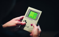 A person is holding a classic handheld gaming device with a green screen displaying a video game. The device appears to be a Game Boy with visible control buttons and a directional pad. The hands gripping the console suggest it is being actively played.