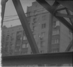 Blurred photograph of a residential building partially obscured by industrial metal beams. The grayscale tones give it a vintage or old film appearance.