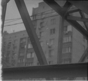 Blurred photograph of a residential building partially obscured by industrial metal beams. The grayscale tones give it a vintage or old film appearance.
