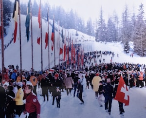 A large crowd of people gathered outdoors on a snowy slope, with numerous colorful flags on tall poles lining the side. The area is surrounded by snow-covered trees, creating a festive and crowded atmosphere.