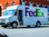 A FedEx Ground delivery truck is parked on a street next to a red brick building. The truck is white with the FedEx logo prominently displayed in purple and green on the side. A driver is seated inside, visible through the open door.