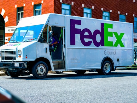 A FedEx Ground delivery truck is parked on a street next to a red brick building. The truck is white with the FedEx logo prominently displayed in purple and green on the side. A driver is seated inside, visible through the open door.