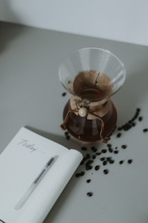 A glass Chemex coffee maker filled with coffee placed on a table next to an open notebook with the word 'Today' written on it. Coffee beans are scattered around the table, and a pen rests on the notebook.