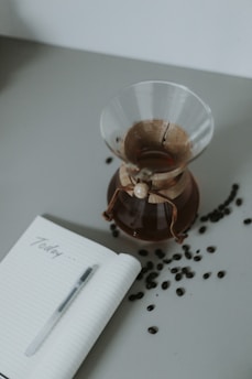 A glass Chemex coffee maker filled with coffee placed on a table next to an open notebook with the word 'Today' written on it. Coffee beans are scattered around the table, and a pen rests on the notebook.