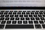 A close-up view of a laptop keyboard featuring a QWERTY layout with black keys and white lettering. Above the keyboard, there is a blurred view of the laptop screen displaying text.
