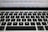 A close-up view of a laptop keyboard featuring a QWERTY layout with black keys and white lettering. Above the keyboard, there is a blurred view of the laptop screen displaying text.