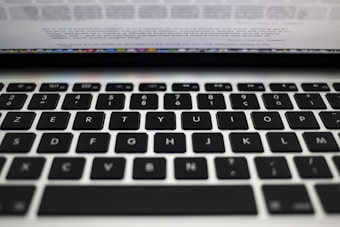 A close-up view of a laptop keyboard featuring a QWERTY layout with black keys and white lettering. Above the keyboard, there is a blurred view of the laptop screen displaying text.