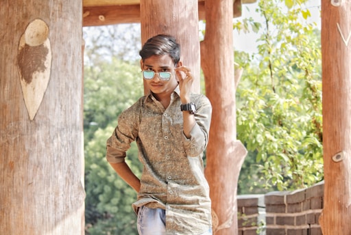 A young person stands confidently with one hand on their hip and the other adjusting sunglasses. The setting includes large wooden beams and lush green foliage in the background, suggesting a natural or outdoor environment.