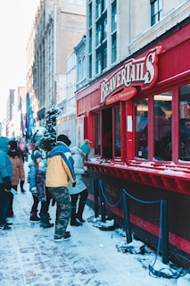 A group of people are standing in line at a food vendor with a bright red exterior. The vendor is located on a snow-covered street in an urban area with tall buildings in the background. Some customers appear to be wearing winter clothing such as jackets and hats, indicating a cold climate.