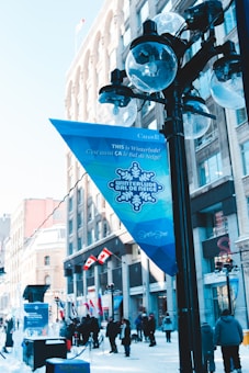 Winter festival scene on a city street featuring a banner that says 'Winterlude' in both English and French. The area is decorated with Canadian flags and includes a few ice sculptures visible on the left. People are gathered around, dressed in winter clothing. Historical buildings line the street, and there is a clear, blue sky overhead.