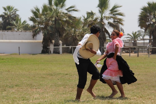 A man and a woman are dancing outdoors on a grassy field. The woman is wearing a traditional pink and black dress with floral accents in her hair, while the man is in dark pants and a white shirt, holding a straw hat. In the background, there are palm trees and white structures.