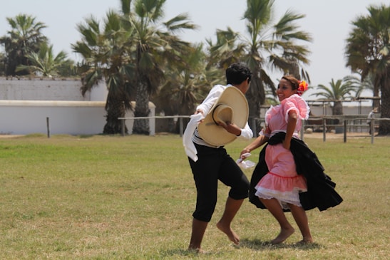A man and a woman are dancing outdoors on a grassy field. The woman is wearing a traditional pink and black dress with floral accents in her hair, while the man is in dark pants and a white shirt, holding a straw hat. In the background, there are palm trees and white structures.