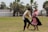 A man and a woman are dancing outdoors on a grassy field. The woman is wearing a traditional pink and black dress with floral accents in her hair, while the man is in dark pants and a white shirt, holding a straw hat. In the background, there are palm trees and white structures.