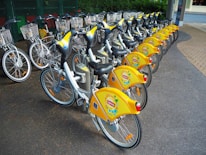 An array of yellow bicycles is neatly arranged in a row at a docking station, ready for use. Each bicycle features a distinctive yellow branding and a wire basket on the front, designed for carrying small items. The setting appears to be an organized bike-sharing system in an urban environment.