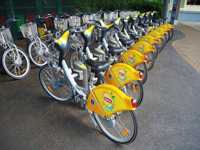 An array of yellow bicycles is neatly arranged in a row at a docking station, ready for use. Each bicycle features a distinctive yellow branding and a wire basket on the front, designed for carrying small items. The setting appears to be an organized bike-sharing system in an urban environment.