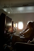 A dimly lit airplane cabin with rows of seats. A woman is sitting in one of the seats, facing forward with light streaming through a window behind her. The overhead compartments and personal entertainment screens are visible.
