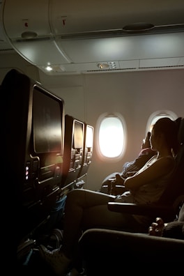 A dimly lit airplane cabin with rows of seats. A woman is sitting in one of the seats, facing forward with light streaming through a window behind her. The overhead compartments and personal entertainment screens are visible.
