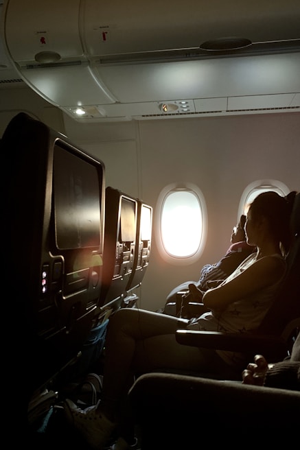 A dimly lit airplane cabin with rows of seats. A woman is sitting in one of the seats, facing forward with light streaming through a window behind her. The overhead compartments and personal entertainment screens are visible.
