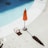A swimming pool with a clear blue surface, partially visible alongside a poolside area. An orange umbrella stands on the concrete deck, casting a shadow. Next to the umbrella, there is a small wooden chair.