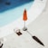 A swimming pool with a clear blue surface, partially visible alongside a poolside area. An orange umbrella stands on the concrete deck, casting a shadow. Next to the umbrella, there is a small wooden chair.