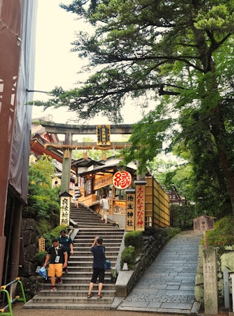 A series of stone steps lead up to a traditional Japanese shrine with wooden signs and lanterns. The path is surrounded by lush greenery, and several people are ascending or photographing the scene. A Torii gate marks the entrance to the shrine, creating a serene and cultural atmosphere.