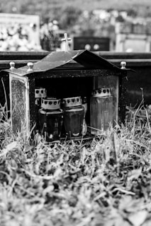 In the foreground, there is a small glass box with a roof and a cross on top, placed on the grass. Inside the box, several candles in glass holders are visible. The background shows a blurred graveyard scene with tombstones and vegetation.