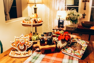 A cozy dining area with warm lighting features a festive Thanksgiving spread. On the table, there is a gingerbread cookie shaped like a turkey, three small bottles of syrup or honey on a wooden stand, a potted red poinsettia plant, and a cookbook open to a colorful page. In the background, there is a two-tiered fruit basket filled with various gourds and oranges. The setting is complemented by an inviting tablecloth and a few decorative lamps, plants, and wall hangings.