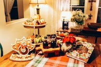 A cozy dining area with warm lighting features a festive Thanksgiving spread. On the table, there is a gingerbread cookie shaped like a turkey, three small bottles of syrup or honey on a wooden stand, a potted red poinsettia plant, and a cookbook open to a colorful page. In the background, there is a two-tiered fruit basket filled with various gourds and oranges. The setting is complemented by an inviting tablecloth and a few decorative lamps, plants, and wall hangings.