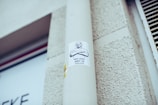 A sticker is attached to a light gray pole, featuring a drawing of a person with crossed arms and a text that reads 'Hey Kids Art is Useless'. The background includes a textured exterior wall and part of a sign with unreadable text.