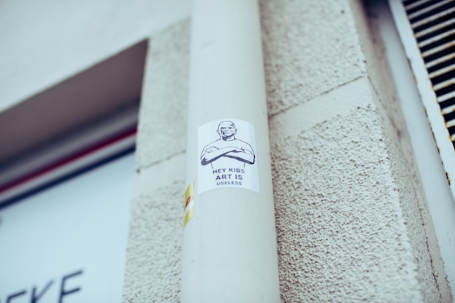 A sticker is attached to a light gray pole, featuring a drawing of a person with crossed arms and a text that reads 'Hey Kids Art is Useless'. The background includes a textured exterior wall and part of a sign with unreadable text.