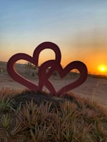 Two intertwined red heart sculptures stand in a sandy, grassy area, with a bright orange sunset on the horizon creating a warm glow across the landscape.