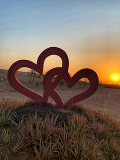 Two intertwined red heart sculptures stand in a sandy, grassy area, with a bright orange sunset on the horizon creating a warm glow across the landscape.