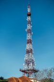 A tall communication tower constructed with a lattice framework rises against a clear blue sky. The tower is equipped with various antennas and satellite dishes. At the base, there are rooftops with terracotta tiles and some sparse tree branches visible.