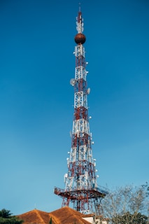 A tall communication tower constructed with a lattice framework rises against a clear blue sky. The tower is equipped with various antennas and satellite dishes. At the base, there are rooftops with terracotta tiles and some sparse tree branches visible.