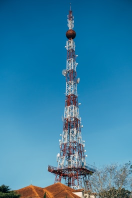 A tall communication tower constructed with a lattice framework rises against a clear blue sky. The tower is equipped with various antennas and satellite dishes. At the base, there are rooftops with terracotta tiles and some sparse tree branches visible.