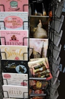 A display rack contains a variety of postcards and greeting cards. The assortment includes cards with cheerful designs, featuring pastel colors, cakes, glitter, and positive messages. Some postcards depict famous landmarks and classic paintings, including a portrait of a woman in an elegant gown. The cards are neatly organized in a metal rack.