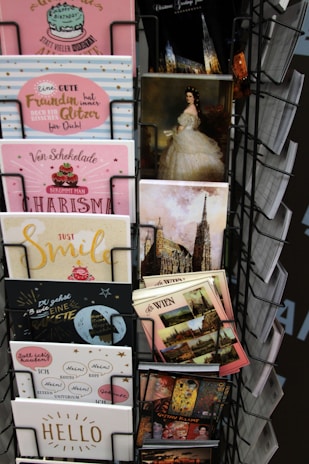 A display rack contains a variety of postcards and greeting cards. The assortment includes cards with cheerful designs, featuring pastel colors, cakes, glitter, and positive messages. Some postcards depict famous landmarks and classic paintings, including a portrait of a woman in an elegant gown. The cards are neatly organized in a metal rack.