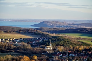 A scenic landscape featuring a village with small houses nestled among lush greenery and a church with a prominent steeple. In the background, there are expanses of open fields and a large, serene body of water surrounded by gentle hills under a pale blue sky.