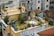 A rooftop garden with numerous potted plants, including green shrubs and small trees, is set on a beige building. A white outdoor table with chairs is positioned prominently in the center, surrounded by various pots and gardening tools. The rooftop also features a small hut with a clay-tiled roof and a large painted mural on the side of the building. Other buildings with balconies and windows are visible in the background, creating an urban setting.