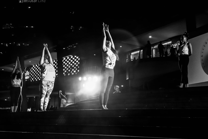 A group of people is practicing yoga or a similar activity on a set of steps in a dimly lit area. The central figures are stretching their arms upwards, while another person nearby is speaking into a microphone. The environment includes architectural elements such as patterned lights and railings, suggesting an urban or public space.