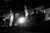 A group of people is practicing yoga or a similar activity on a set of steps in a dimly lit area. The central figures are stretching their arms upwards, while another person nearby is speaking into a microphone. The environment includes architectural elements such as patterned lights and railings, suggesting an urban or public space.